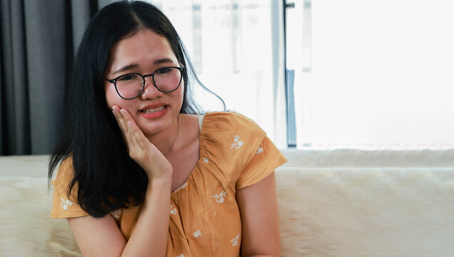 Young Asian woman was experiencing oral health problems specifically tooth decay displayed pained expression suffering from severe toothache jaw pain and headache simultaneously.
