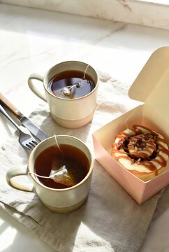 Two cups of hot tea with teabags served with cinnamon roll in pastel box on soft linen cloth.