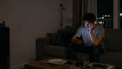 Obraz premium Man sitting on sofa at night looking at smartphone, feeling lonely in dimly lit living room