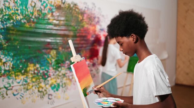 Closeup of african boy hand paint canvas with warm tone color while caucasian girl in casual outfit drawing cool tone colors behind at colorful stained wall. Creative activity concept. Edification