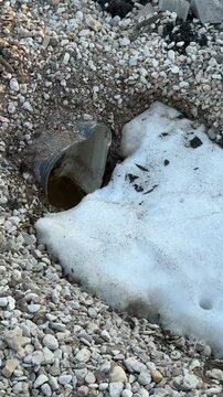 Snow crushed stone around sewer pipe, sunlit urban curb and asphalt, melting patches and scattered pebbles, exposed drain hole