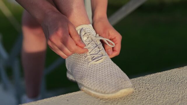 A person is kneeling to tie shoelaces on a pair of light-colored shoes. The setting is sunny and green, perfect for outdoor exercise