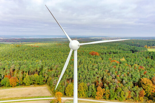 Simmerath, Germany - 09 May 2026: Aerial view of a large white wind turbine standing tall over a dense forest with autumn foliage under a cloudy sky.