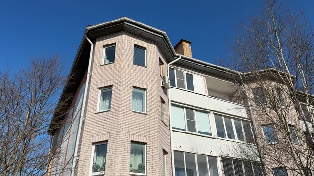 White brick apartment against blue sky. Sunlit multistorey facade with rows of windows, balconies and chimney framed by bare