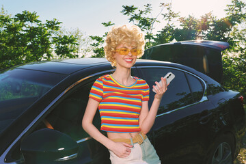 Fototapeta premium Smiling woman with curly hair wearing a striped croptop stands beside a sleek car on a sunny day, expressing a vibrant lifestyle and retro fashion mood. Soft daylight enhances warm tones, suggesting