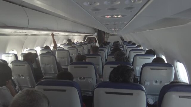 Passengers in airplane seats, looking forward and waiting for departure or during flight, with overhead compartments visible.