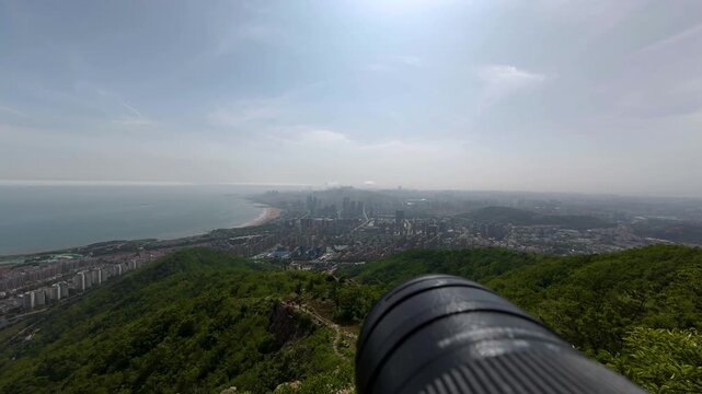A high-angle panoramic shot taken from a mountain summit, showcasing a coastal city, adjacent beach, forested hills, and a clear daytime sky with a viewing telescope in the foreground.