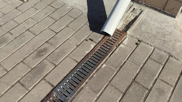 White downpipe and metal grate on paving stones, sunlight casting shadow across textured pavers, closeup view of storm water