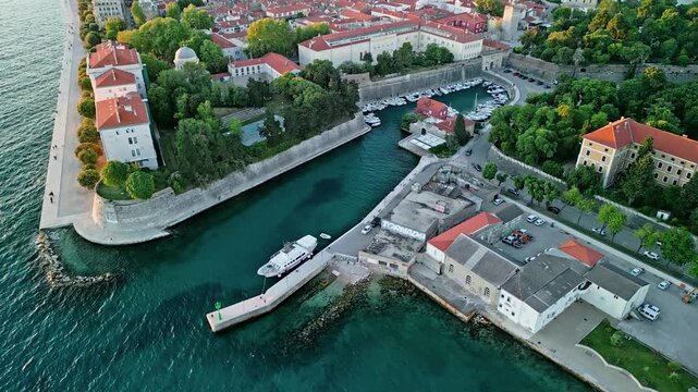 Zadar, Croatia - 05 17 2025: Dolly drone video moving backward showing Molo Terraferma Marina, turquoise Adriatic Sea, harbor with boats and docked ferry, historic buildings, Land Gate, lush greenery