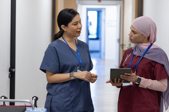 Two female medical workers in scrubs standing in hospital corridor discussing tablet, stethoscope