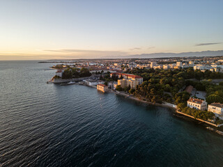 Obraz premium Zadar, Croatia - 05 17 2025: Angled drone photo of Zadar coastline at sunset, showing Adriatic Sea on the left, city buildings on the right, Vladmir Nazor’s Grammar School, mountains in the distance