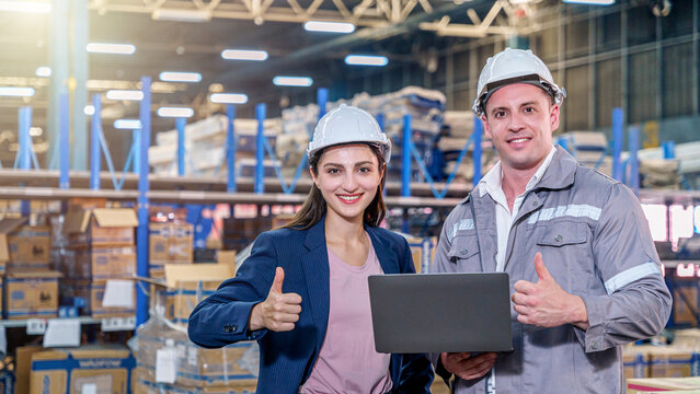 A smiling female manager in a blue blazer and a male worker in grey coveralls stand side-by-side in a large, brightly lit warehouse. Both are wearing white safety hard hats. Giving thumbs-up gesture.