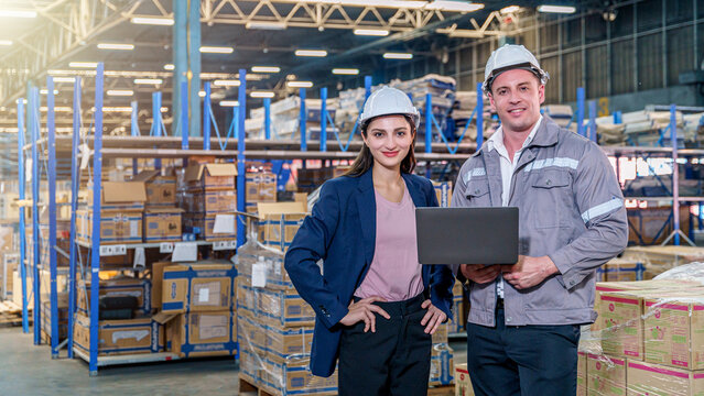 A smiling female manager in a blue blazer and a male worker in grey coveralls, both wearing white hard hats, stand confidently inside a busy, well-lit warehouse. The man holds an open laptop.