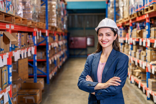 A smiling female supervisor stands confidently with her arms crossed in the aisle of a large warehouse. She is wearing a dark blue pinstripe blazer over a pink top and a white safety hard hat. The bac