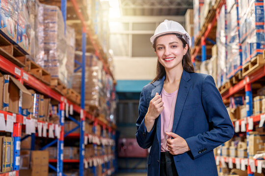 A smiling female supervisor stands confidently. She is wearing a dark blue pinstripe blazer over a pink top and a white safety hard hat. The background features tall, orange and blue industrial storag