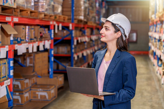 A female warehouse supervisor in a dark blue pinstripe blazer and a white safety hard hat stands in a storage aisle, looking up toward the shelving racks. She holds an open laptop in her hands, conduc