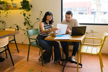Fototapeta premium Two diverse colleagues reviewing paperwork and collaborating on a new work task at a casual coffee shop