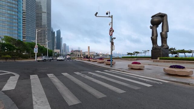 This is an overcast daytime urban coastal street scene, with marked crosswalks, moving vehicles, modern bronze sculptures, lined skyscrapers and roadside flower beds.