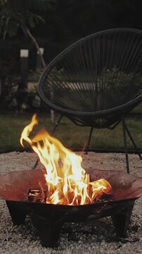 Vertical video. Burning firewood in metal fire bowl in backyard during summer evening outdoors. Warm flames and glowing embers create relaxing atmosphere. Filmed in 4K slow motion.