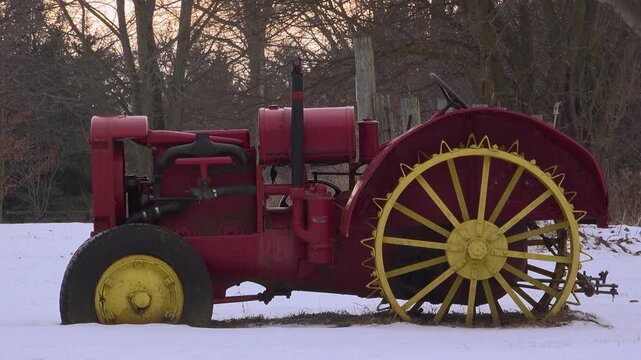 A side profile shot of a classic antique red tractor featuring large yellow steel cleated wheels and a rubber front tire, parked in a snow-covered farm field with winter trees behind it at sunset.