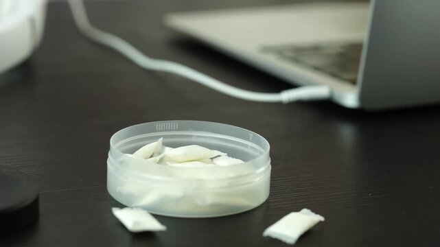 an open round plastic container with white nicotine pouches and snus pads on a dark wooden desk setup with white overhead headphones and a blurry laptop in the background. 