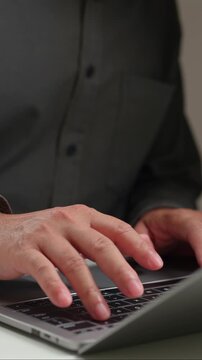 Male worker wearing dark shirt is typing code, inputting data inside quiet office workspace as fingers pressing keys operate laptop keyboard, closeup view showing hands performing work tasks.