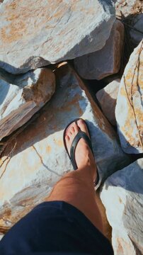 Person's feet in flip-flops stand on sunlit rocks. Sunlight highlights rough stone textures. View shows hiking or exploring rocky terrain. Footage captures outdoor adventure and casual travel vibes