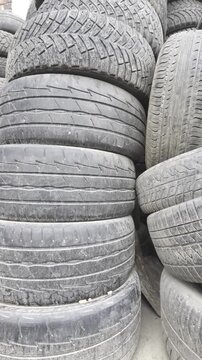A large stack of old used car tires lies piled up outdoors. The worn rubber protectors and tread patterns are covered in dust and dirt.