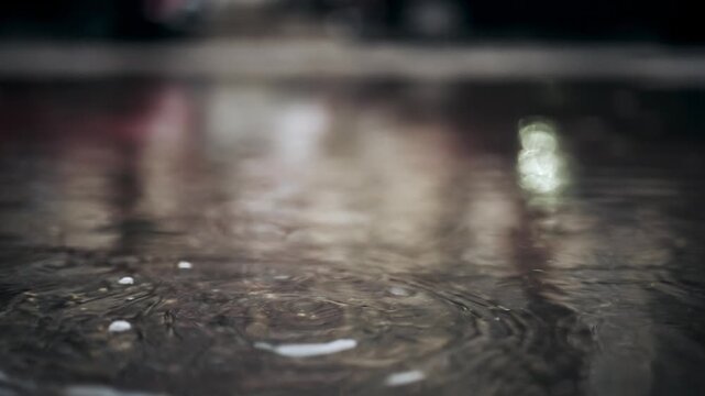 Rain drops falling into puddle on asphalt surface during heavy downpour.