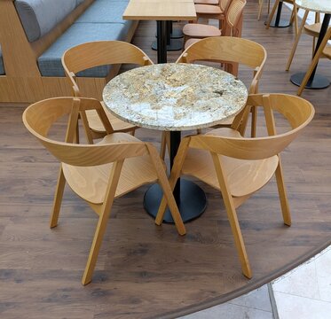 Primer plano de una mesa redonda con cuatro sillas de madera, con fondo de mesa cuadrada con sillas de madera y un asiento de banco con cojines, en terraza de un restaurante de un centro comercial	