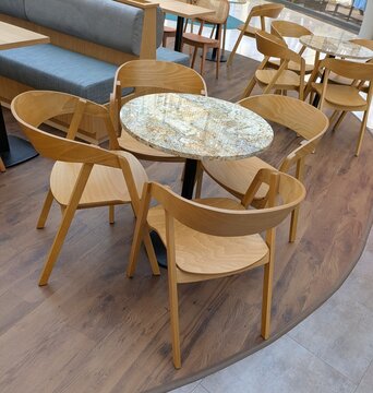 Primer plano de una mesa redonda con cuatro sillas de madera, con fondo de mesa cuadrada con sillas de madera y un asiento de banco con cojines, en terraza de un restaurante de un centro comercial	