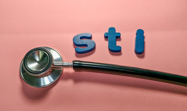 Close-up of a stethoscope placed on a pastel pink background beside blue wooden letters spelling &ldquo;STI.&rdquo; The minimalist composition symbolizes sexual health and public health awareness.