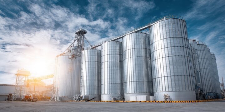 Industrial farm agricultural silos for storage and drying of wheat grains, sugar, corn, soy, sunflower on blue sky. Large metal grain tanks processing equipment in modern facility. Agriculture, food.