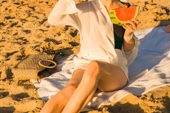 Enjoying watermelon and drinks on the beach during a sunny day with friends while sitting on a blanket.  woman sits on sandy beach. girl holds  and eats watermelon slices .  clear, sunny day.