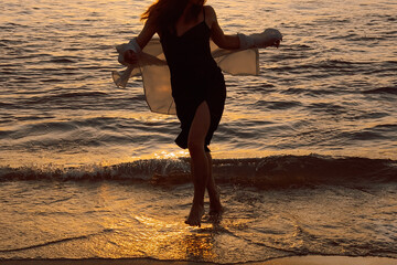 Silhouette  person with arms outstretched at beach during sunset near ocean waves. woman stands at edge  ocean with arms raised, looking towards sunset. sky shows warm colors as waves © SValeriia