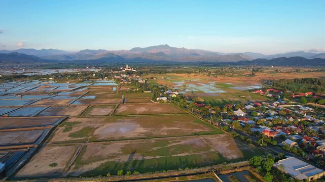 Aerial drone footage of a dry rice field valley, with villages in Woha area, Sumbawa, Indonesia, at golden sunset, salt farm ponds, a main road, and Agung main mosque of Bima regency, mountains behind