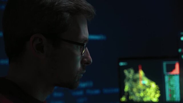 Close-up of focused data scientist in eyeglasses in a dark office writing code on keyboard and appears deeply concentrated, working late on a complex cybersecurity or AI programming task. 