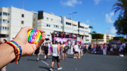 Obraz premium Human hand showing a rainbow love is love badge and wristband, celebrating equality and diversity at an LGBTQ+ pride parade with a blurry crowd in the background