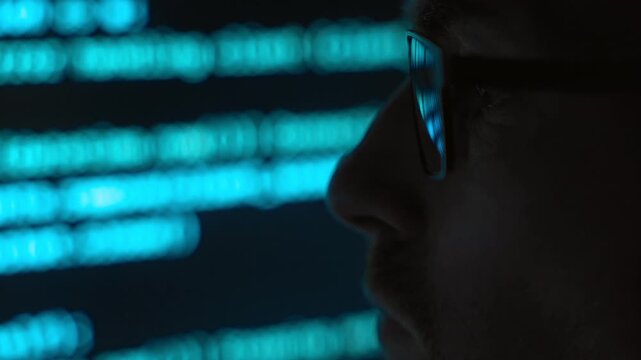 Side view close up, focused software engineer in eyeglasses looking at a blue code running on screen in a dark room, programming script reflects in his lenses. AI and data science concept. Copy space
