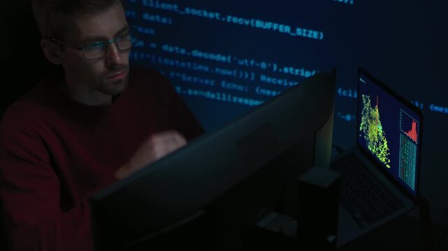 Software engineer in glasses sits in a dark room at a desk with multiple computers. Thoughtful male developer works on complex data science project, programming script lines moving on a wall behind.