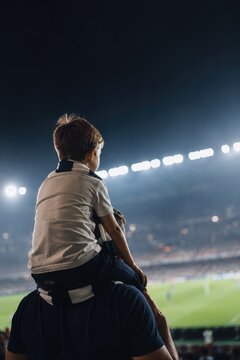 Little boy sitting on shoulder of father watching soccer match from stadium stand. Child and man athlete fan at game. Family leisure time during night championship competition event