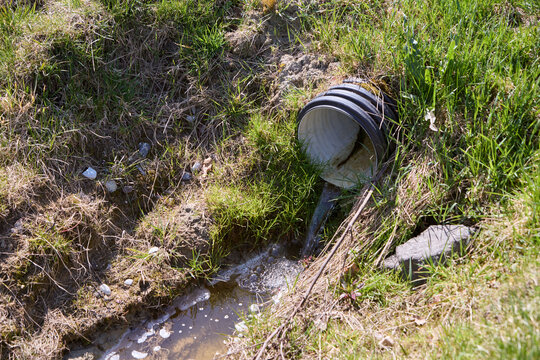 Black plastic drainage pipe discharging water onto grassy slope under sunlight
