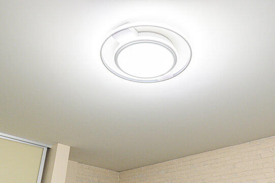 sleek white circular LED ceiling light fixture with concentric rings is mounted on a smooth off-white ceiling, casting a soft glow over a light brick wall