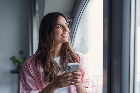 Young adult woman holding a smartphone, smiling and looking away through a window, reflecting optimism, connection, and a hopeful future in an indoor setting