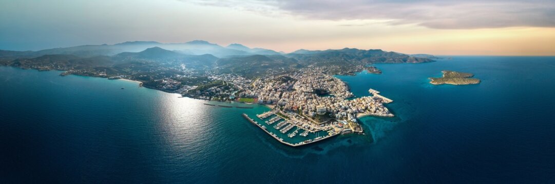 Aerial panorama 17k of Agios Nikolaos city and Lake Voulismeni in Crete Geece. Drone view yacht marina, circular lake and scenic coastline of Mirabello Bay at golden hour. Urban landscape