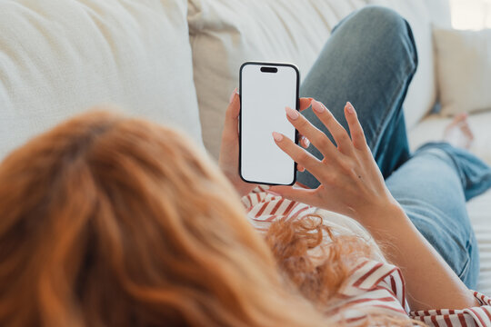 Red-haired woman relaxing on a cozy couch, holding a smartphone with a blank white screen and tapping it with her finger, perfect for app, social or advertising mockups
