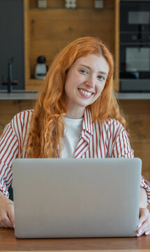 Smiling woman with red hair looking at the camera while working remotely on her laptop, depicting modern work flexibility, career success, and a connected lifestyle