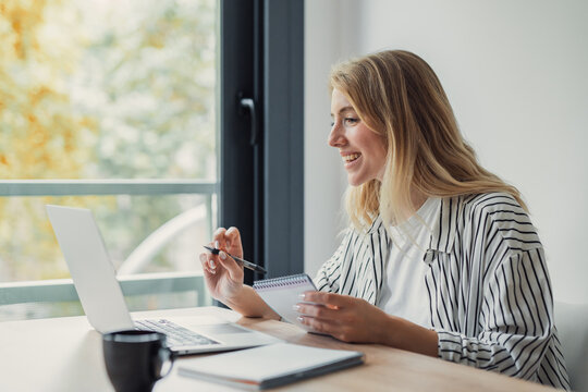 Young woman smiling at camera while taking notes during a laptop video call in a bright home office, studying or working remotely as a cheerful freelancer or student