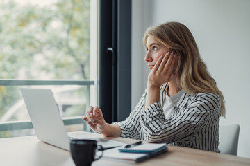 Obraz premium Young woman looking away from her laptop, sitting at a desk and contemplating with her hand resting on her face, possibly taking a break from remote work