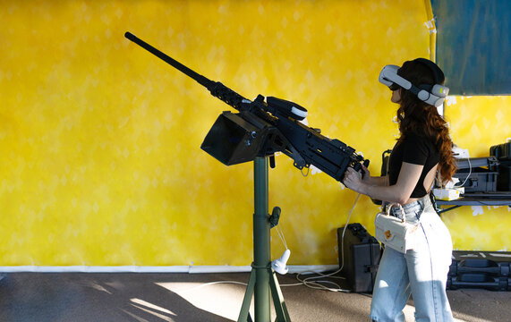 Young ukrainian girl wearing virtual reality glasses playing with heavy machine gun aims to shoot down a Shahed attack drone. Arms and Safety Exhibition. 